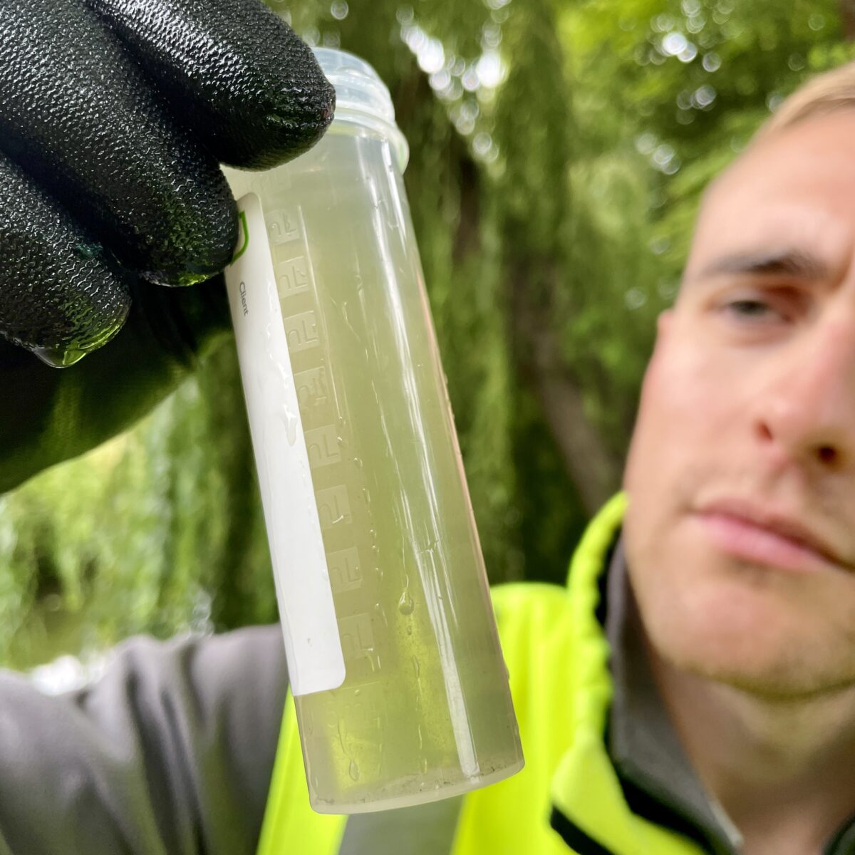 A technician wearing gloves and a hi vis jacket inspects a test tube of water.