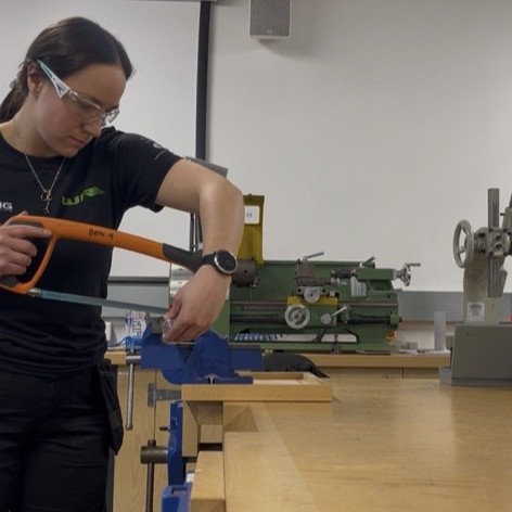 Bethany in the workshop, wearing safety goggles, using a saw.