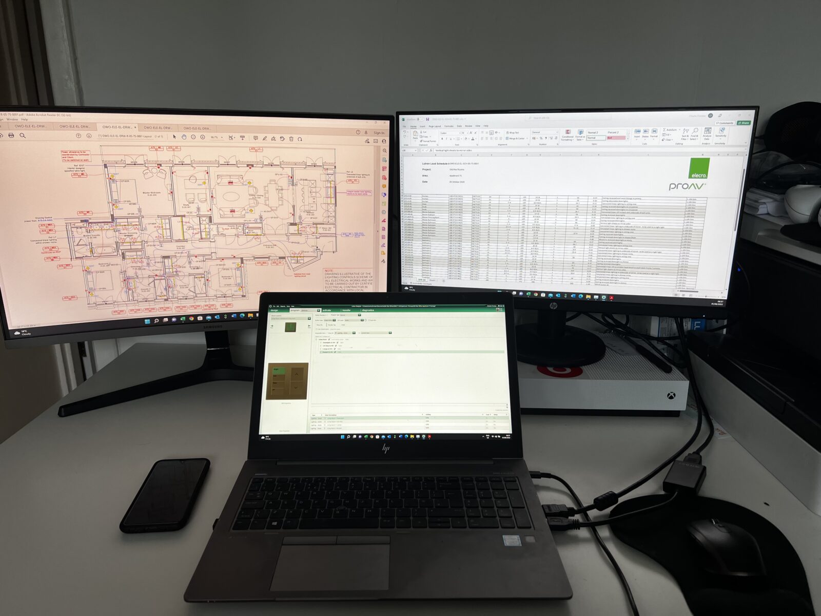 Charlie Finniear's workstation, a desk with a laptop and two screens, showing spreadsheets and building plans.