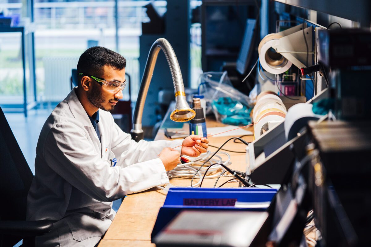 A male battery technician, wearing a lab coat and goggles, works at a desk covered with electrical equipment.