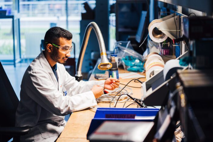 A male battery technician, wearing a lab coat and goggles, works at a desk covered with electrical equipment.