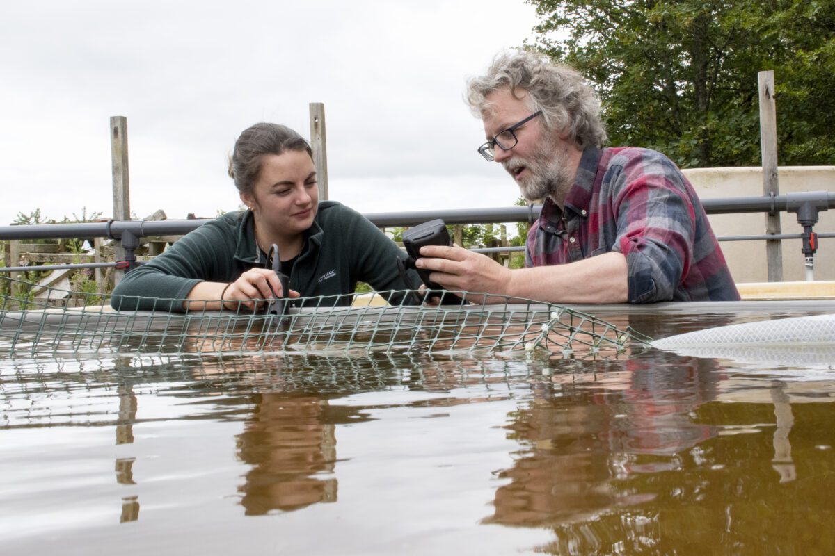Two aquaculture technicians, a man and young woman, next to a pool of water, taking a water reading.