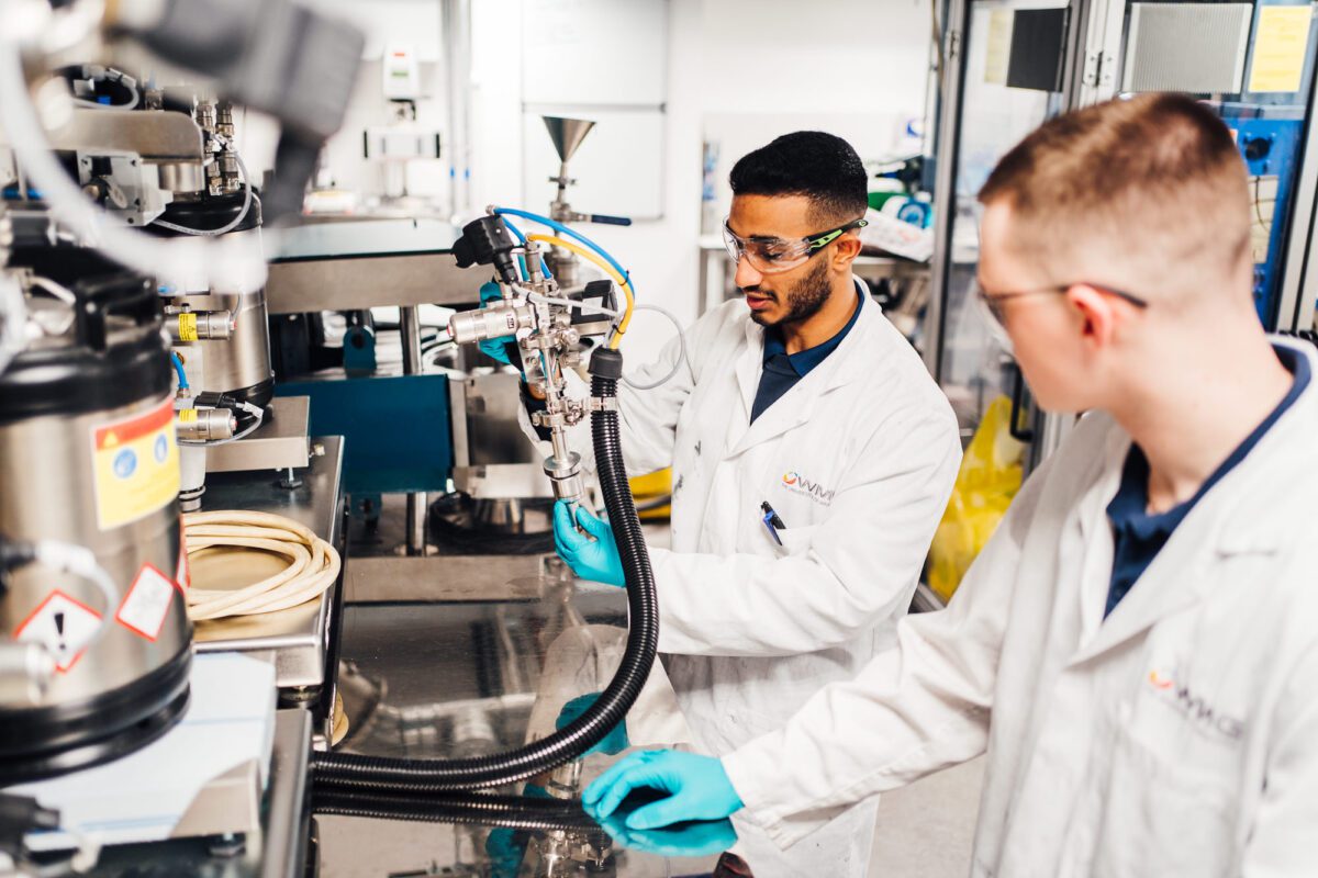 Divesy and a colleague using industrial equipment, wearing lab coats, safety glasses, and gloves.