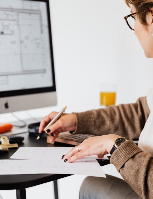Woman at desk taking notes from a screen showing building plans.