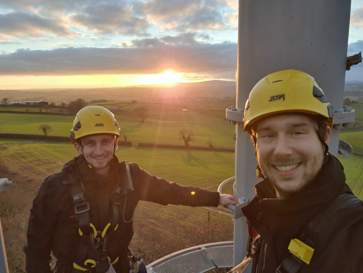 Lewis taking a selfie, smiling with a co-worker at the top of a telephone mast, surrounded by a rural landscape.