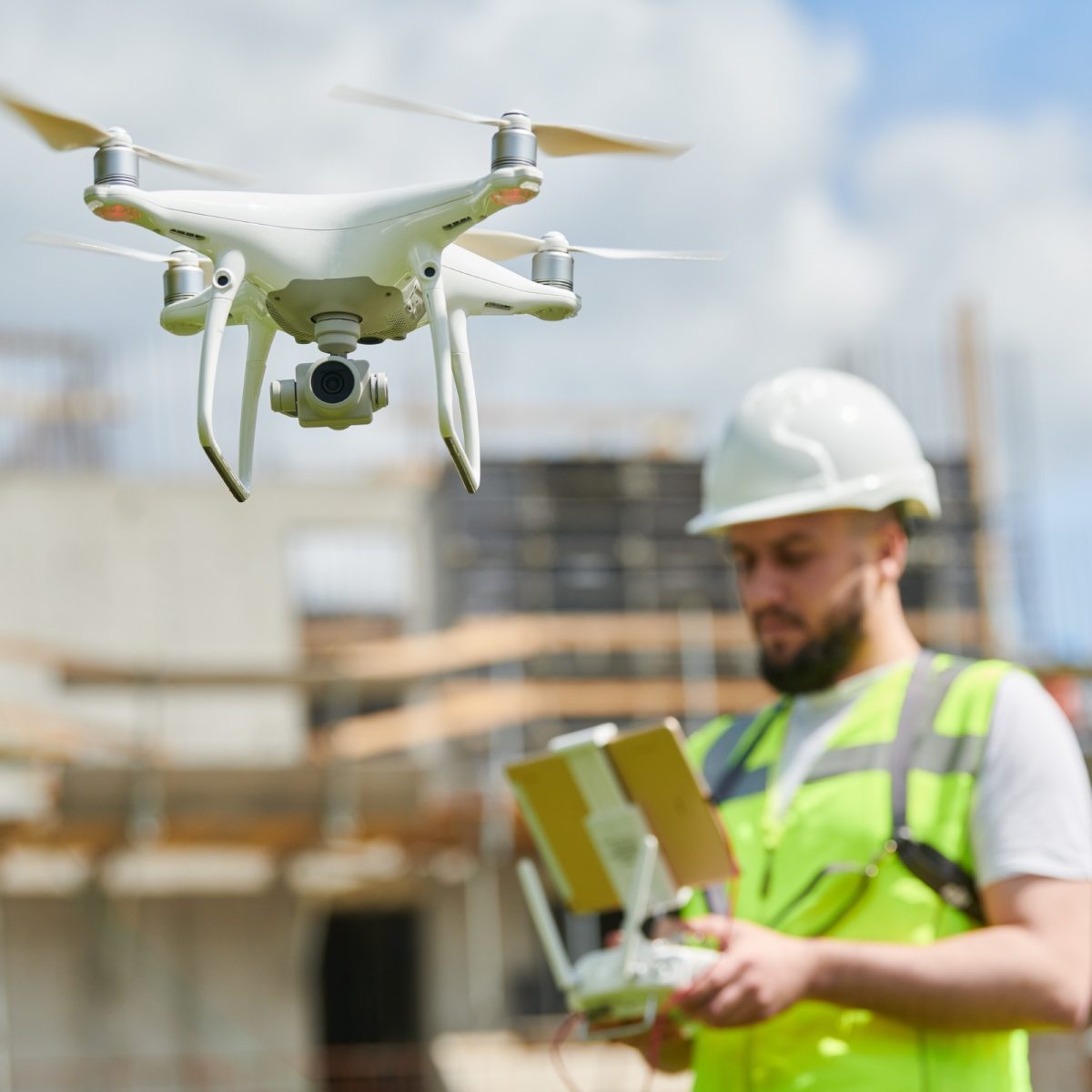 A person on a construction site in a hi-vis jacket and hard hat, flying a drone by remote.
