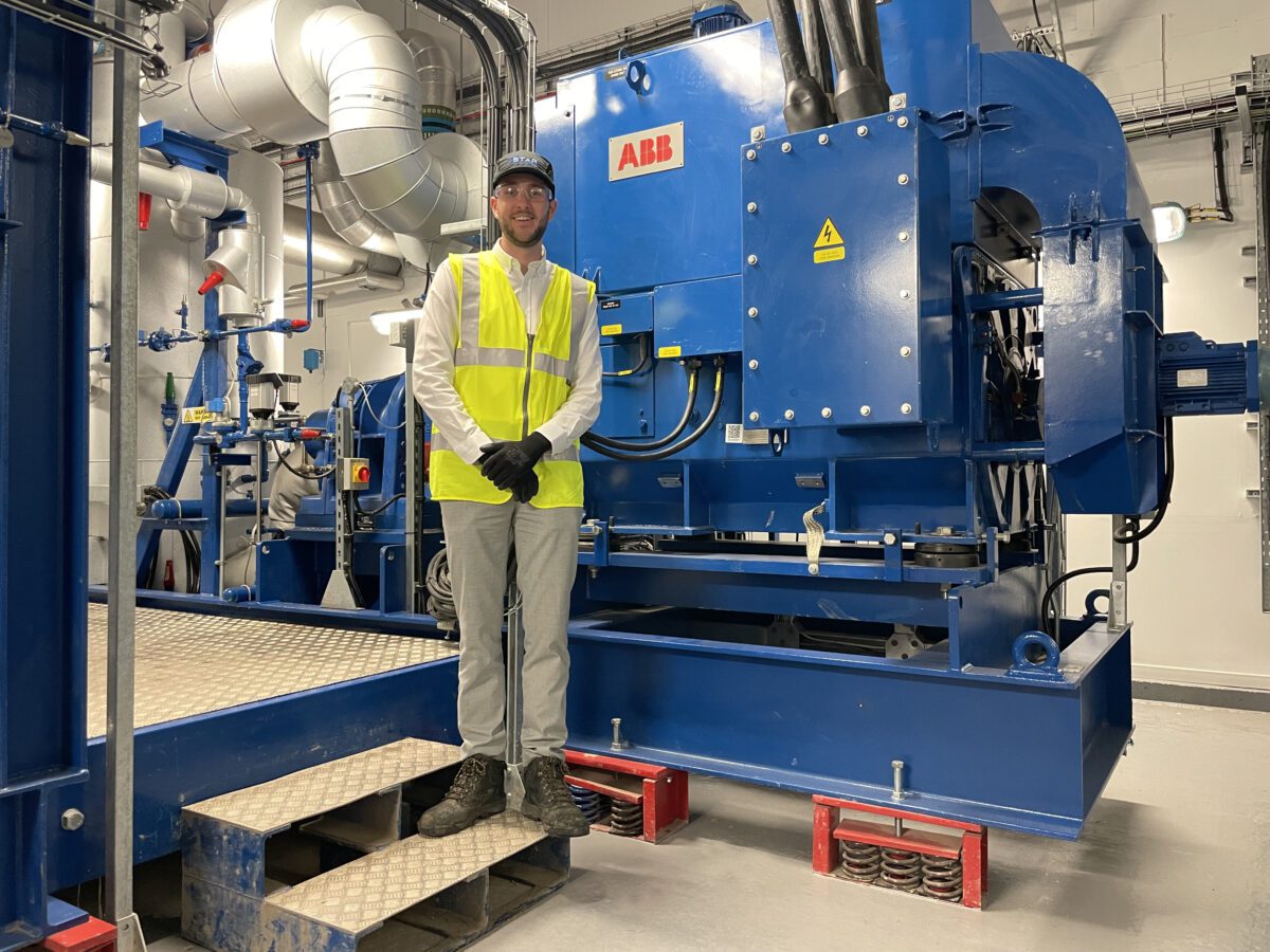 Tom at work, posing in front of a large industrial machine, wearing a hi-vis jacket and safety glasses.