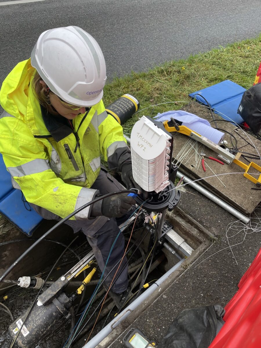 Chloe Williams working on underground broadband cables next to a road, wearing a hi-vis jacket, hard hat, gloves, and safety goggles.