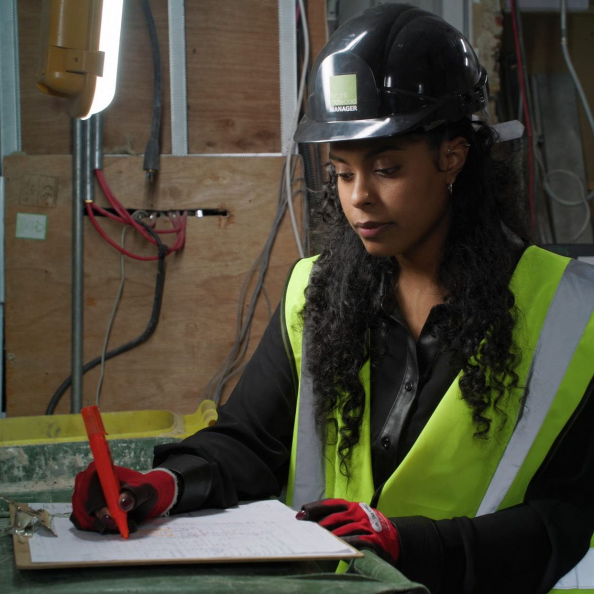 Building Services Technician Leteesha, working in a construction site, wearing a hi-vis vest, hard hat and gloves, marking up some drawings on a clipboard.