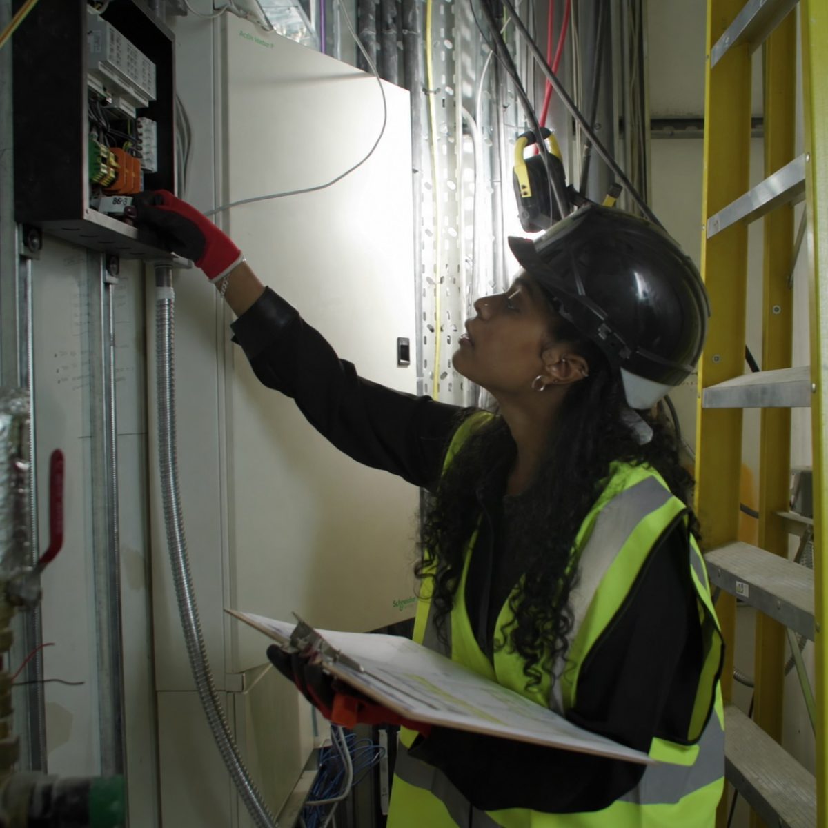 Building Services Technician Leteesha, working in a construction site, wearing a hi-vis vest, hard hat and gloves, checking an electrical circuit breaker box.