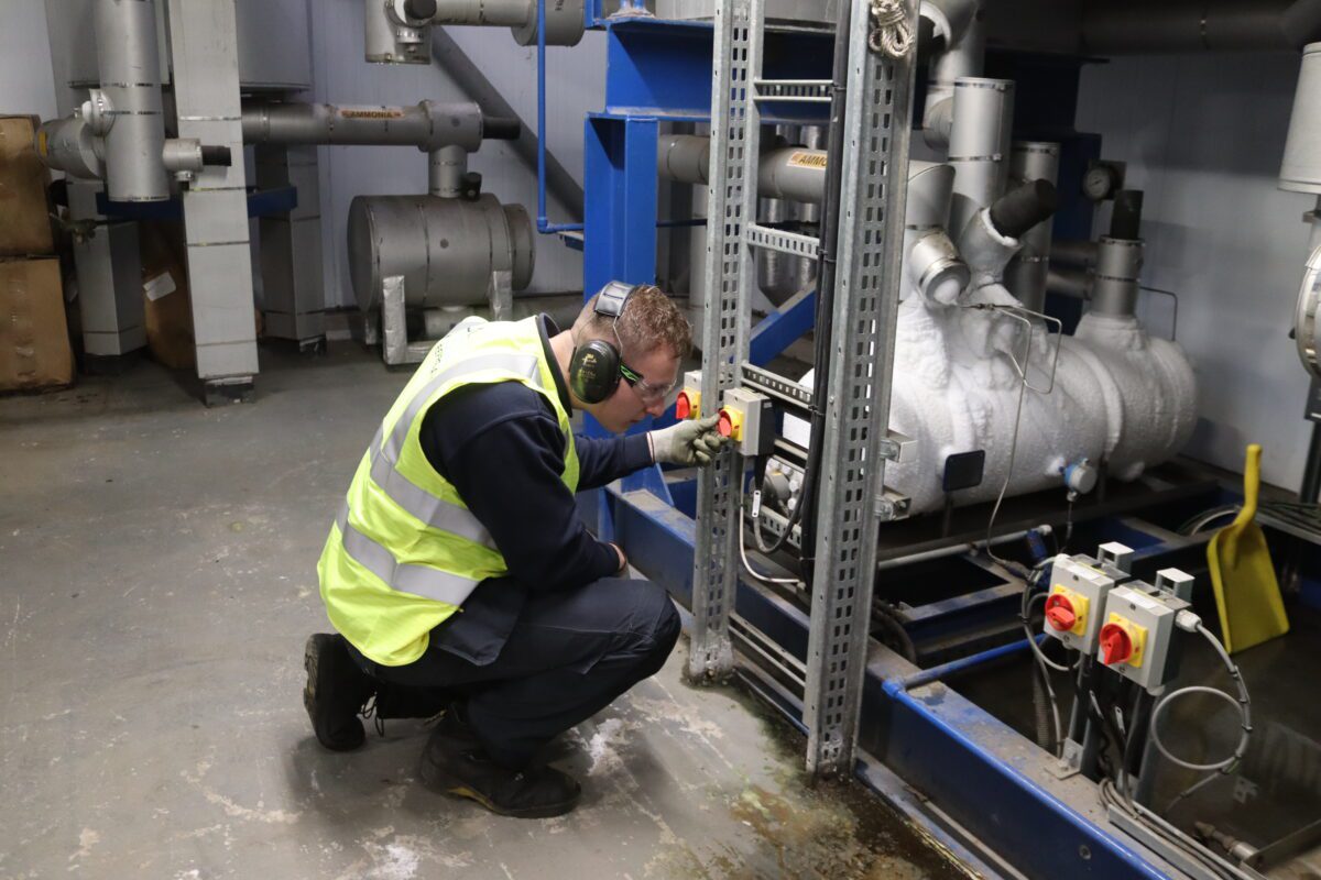 Sam kneeling to check and test a piece of industrial refrigeration equipment, whilst wearing a hi-vis jacket, safety glasses, ear protectors, and gloves.