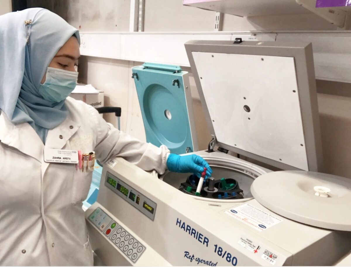 Zahra at work in the lab, placing samples in a centrifuge, wearing a lab coat and protective gloves.