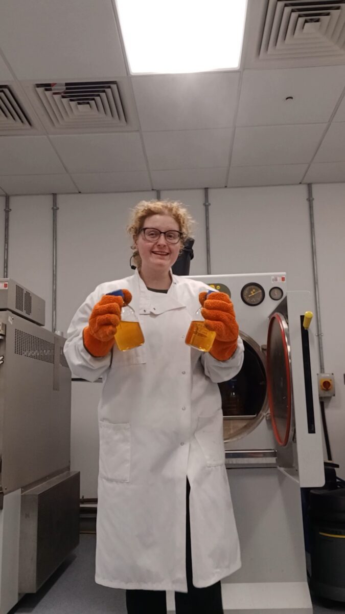River holding two bottles of liquid in the lab, wearing a lab coat, and large protective gloves.