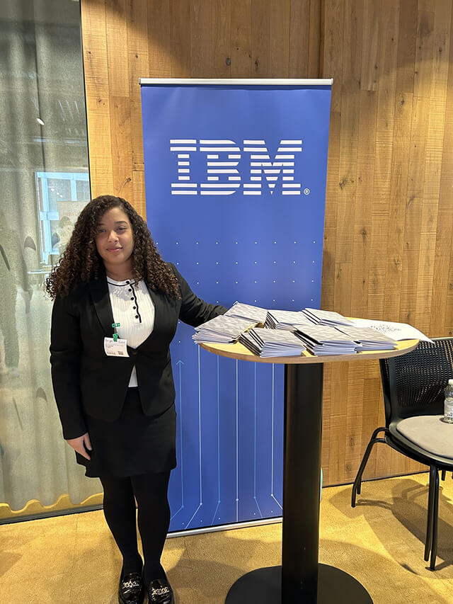 Ferndanda standing next to an IBM branded banner and table, ready to speak to people at a networking event.