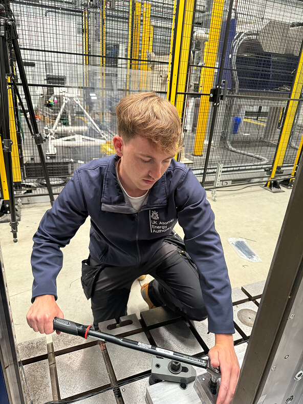 Sam using a hand tool to fix a piece of equipment, on a workshop floor, wearing overalls.
