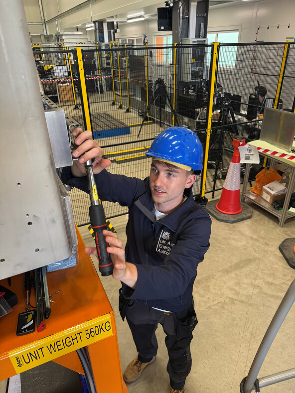 Sam using a torque wrench in a workshop, wearing a hard hat and overalls.