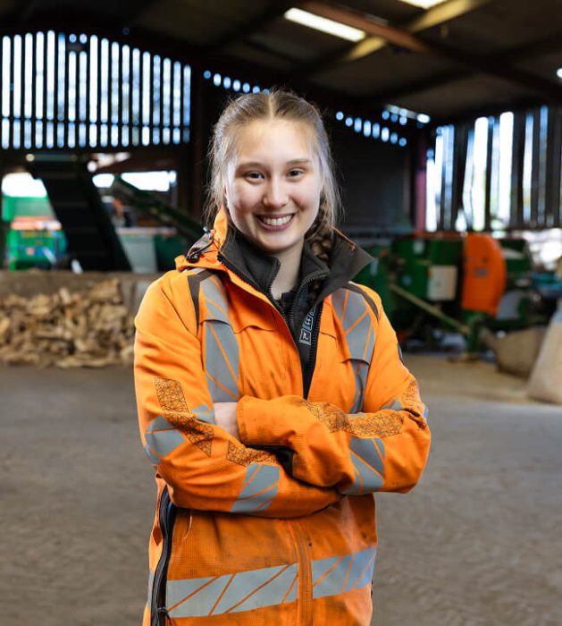 A picture of a woman in an orange high vis jacket with her arms folded facing the camera smiling.