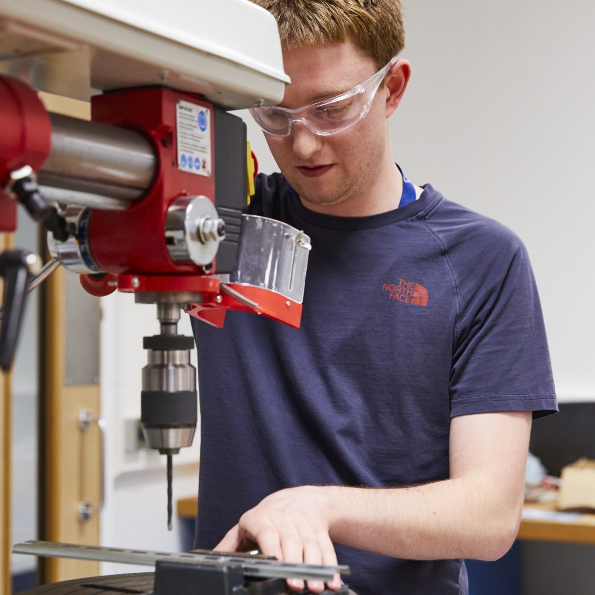 Louis, wearing protective glasses, using a drill press to drill a hole in a piece of metal.