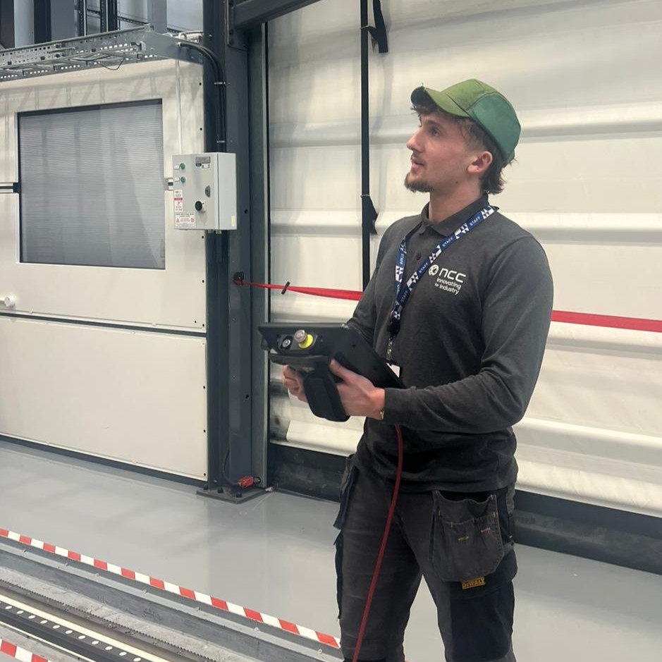 Sam wearing a baseball cap, in the workshop, controlling a piece of machinery with a tablet.