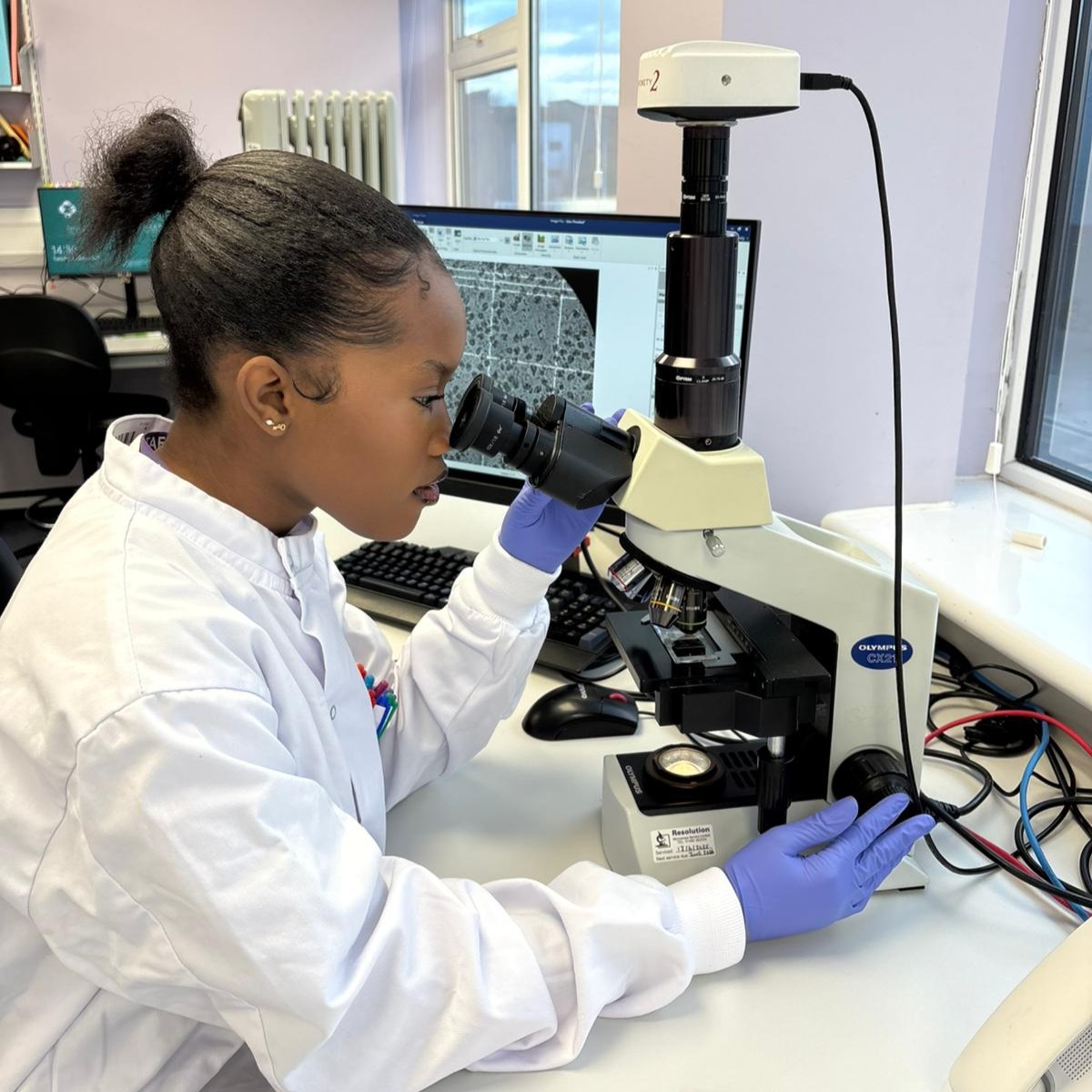 Dudzai in the lab, wearing a lab coat and a pair of nitrile gloves, looking through a microscope at some cells.
