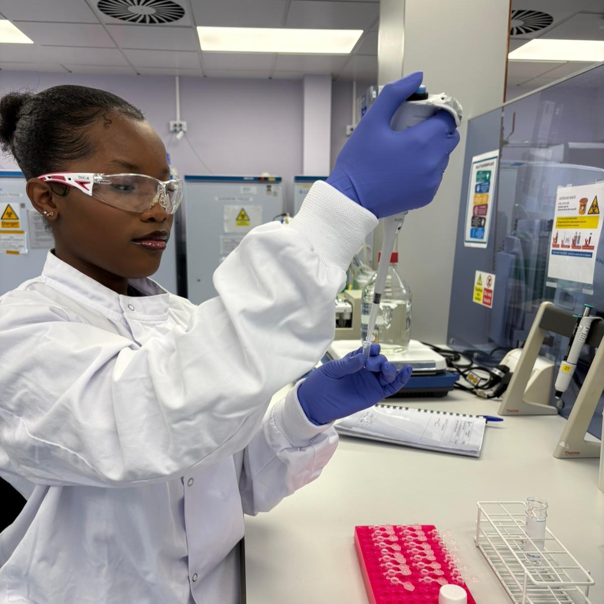 Dudzai in the lab, wearing a lab coat, safety goggles, and a pair of nitrile gloves, using a micropipette to squirt liquid into a small test tube.