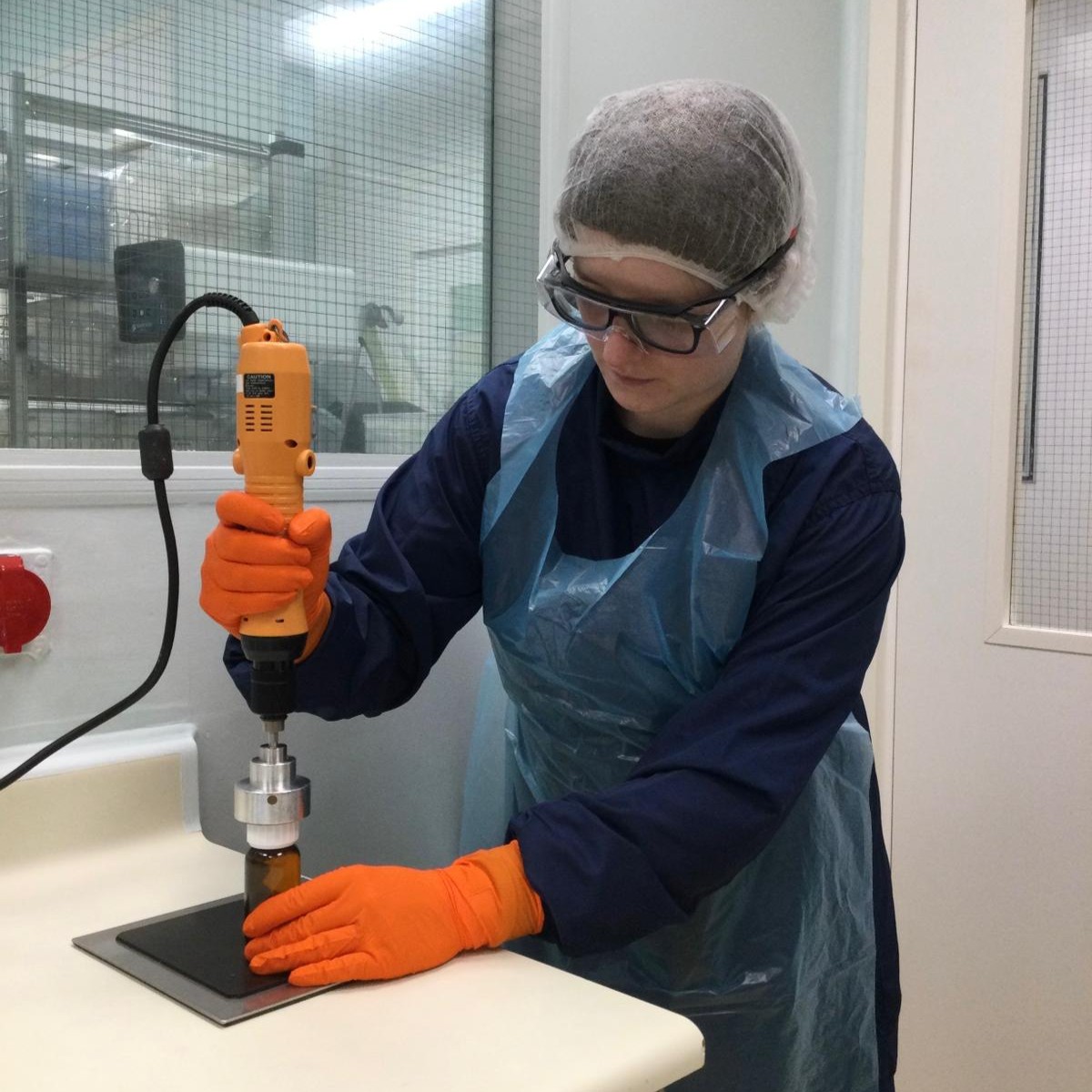 Louisa wearing an apron, hair net, safety goggles, and gloves, mixing medicines with an electric mixer in the lab.