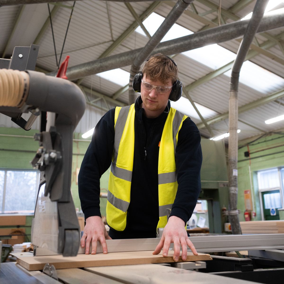 Will wearing a hi-vis jacket, safety goggles, and ear defenders, cutting a piece of wood in the joinery workshop.