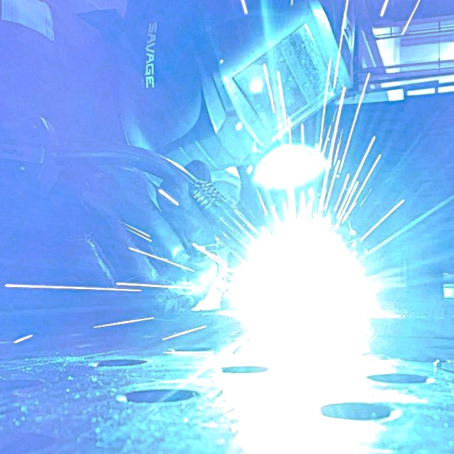 A close-up of Lily welding metal in a welders mask, with hot plasma and sparks.