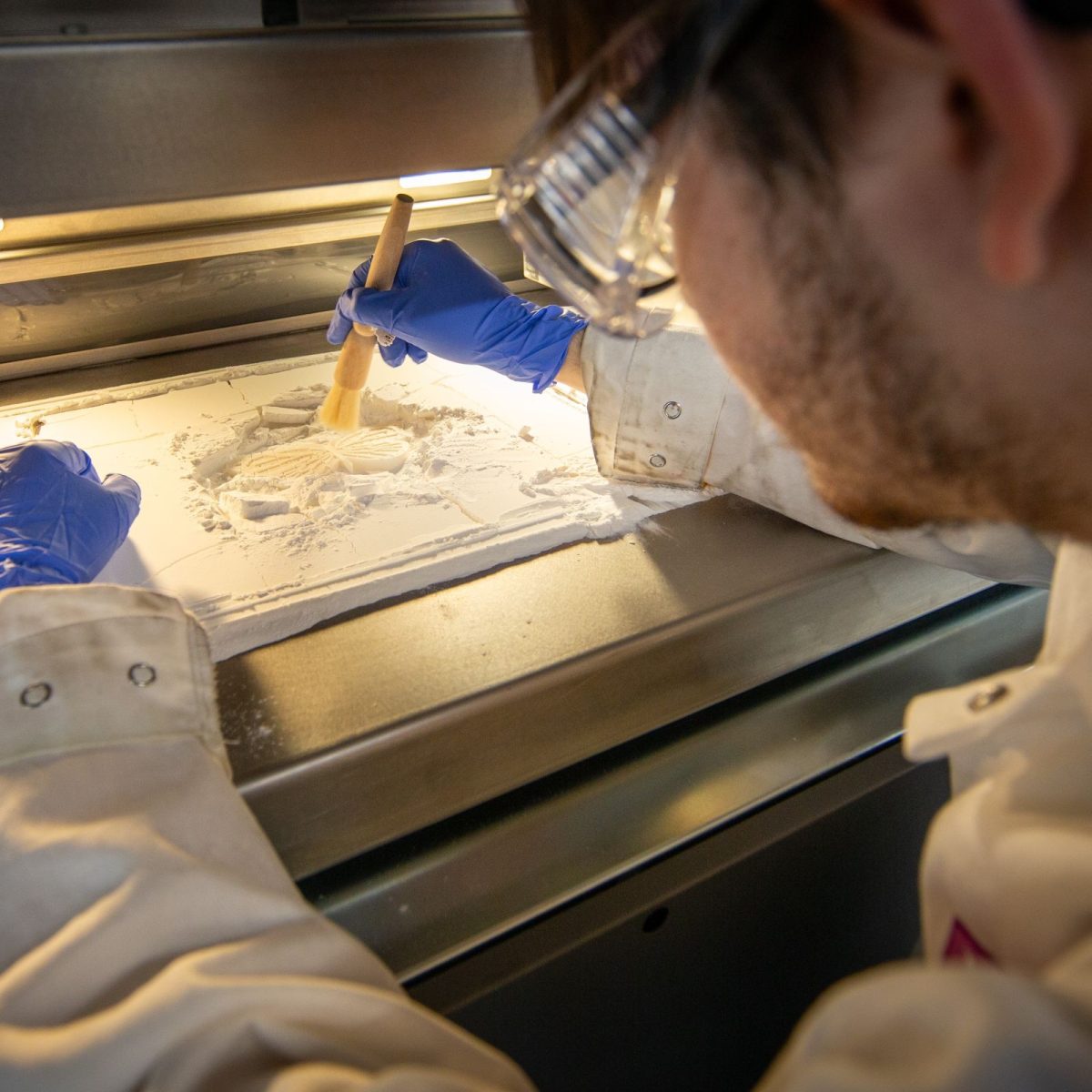 Olly, wearing a lab coat, gloves, and safety glasses, mixing a powder with a brush, in the lab.