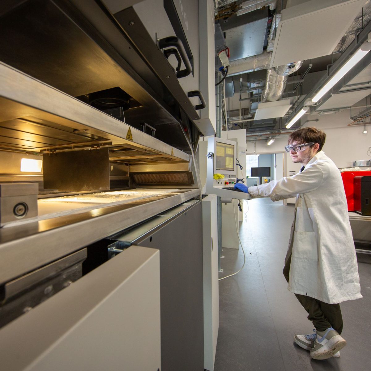 Olly, wearing a lab coat, gloves, and safety glasses, using a large machine in the lab.