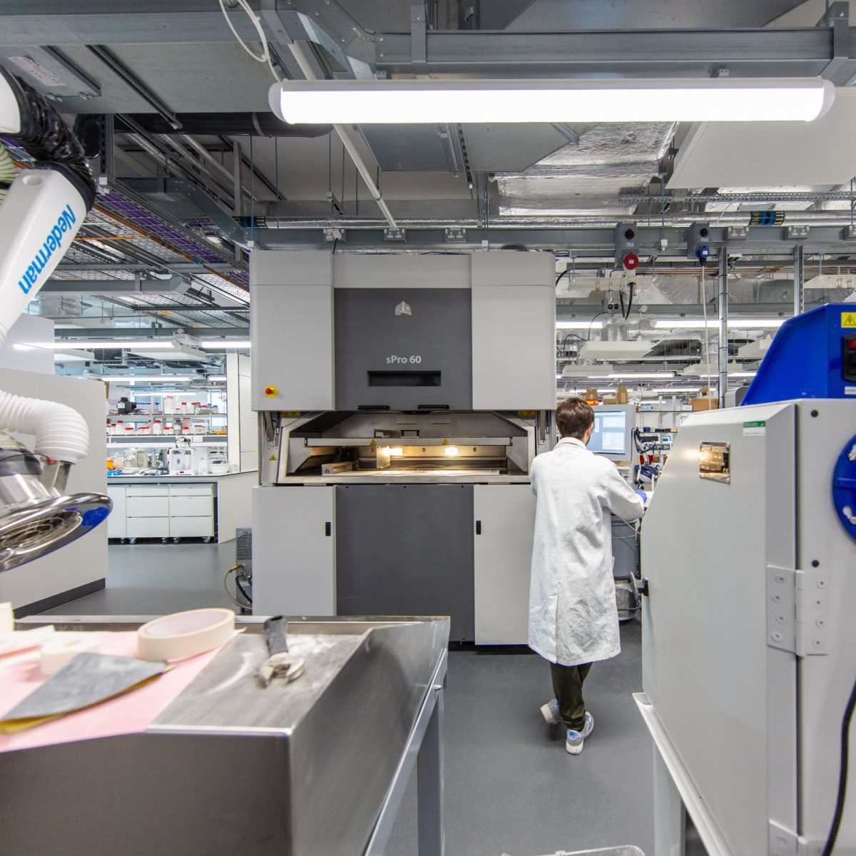 Olly, wearing a lab coat, gloves, and safety glasses, surrounded by large machines in the lab.