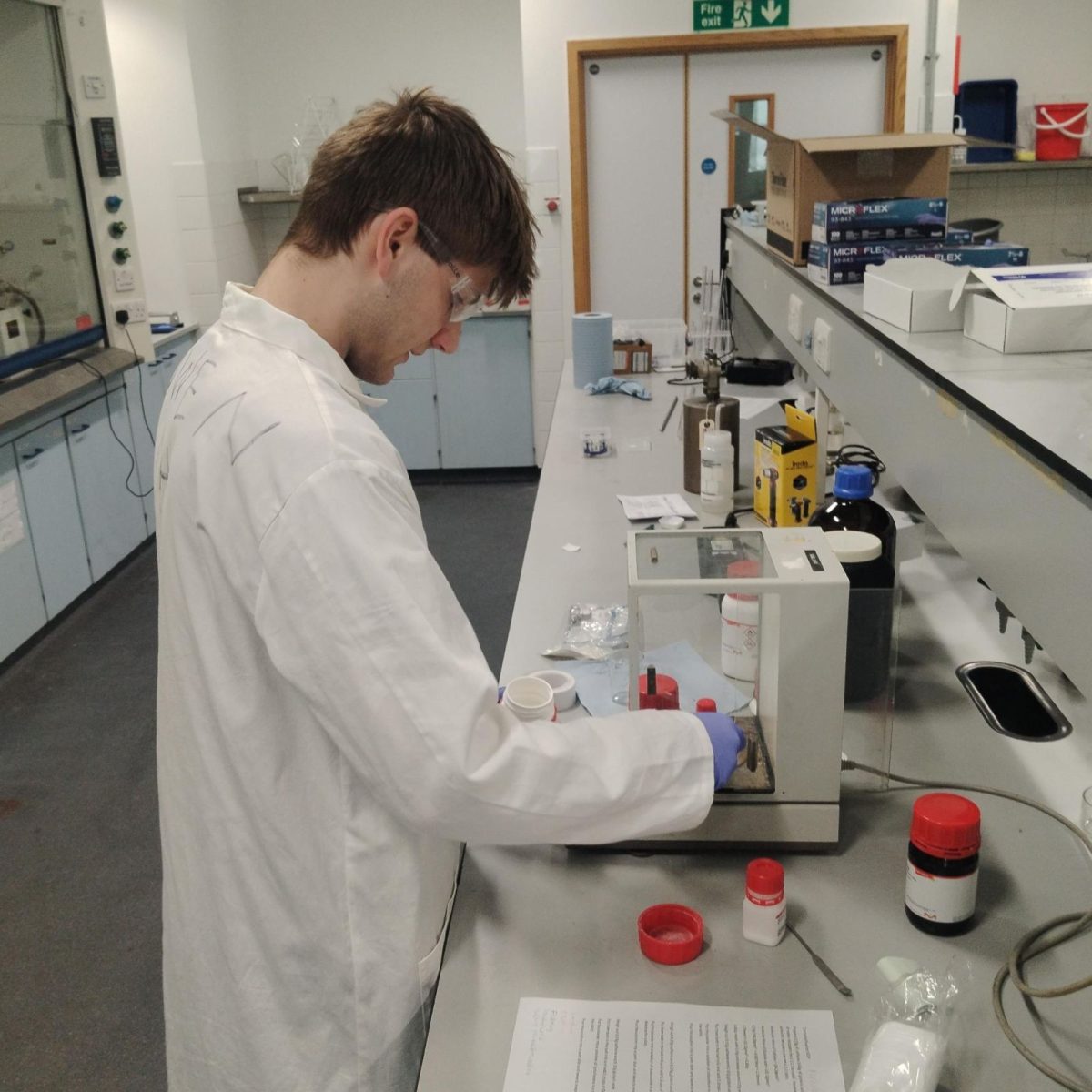 Owen in the lab, wearing a lab coat, gloves, and safety goggles, measuring out some samples.