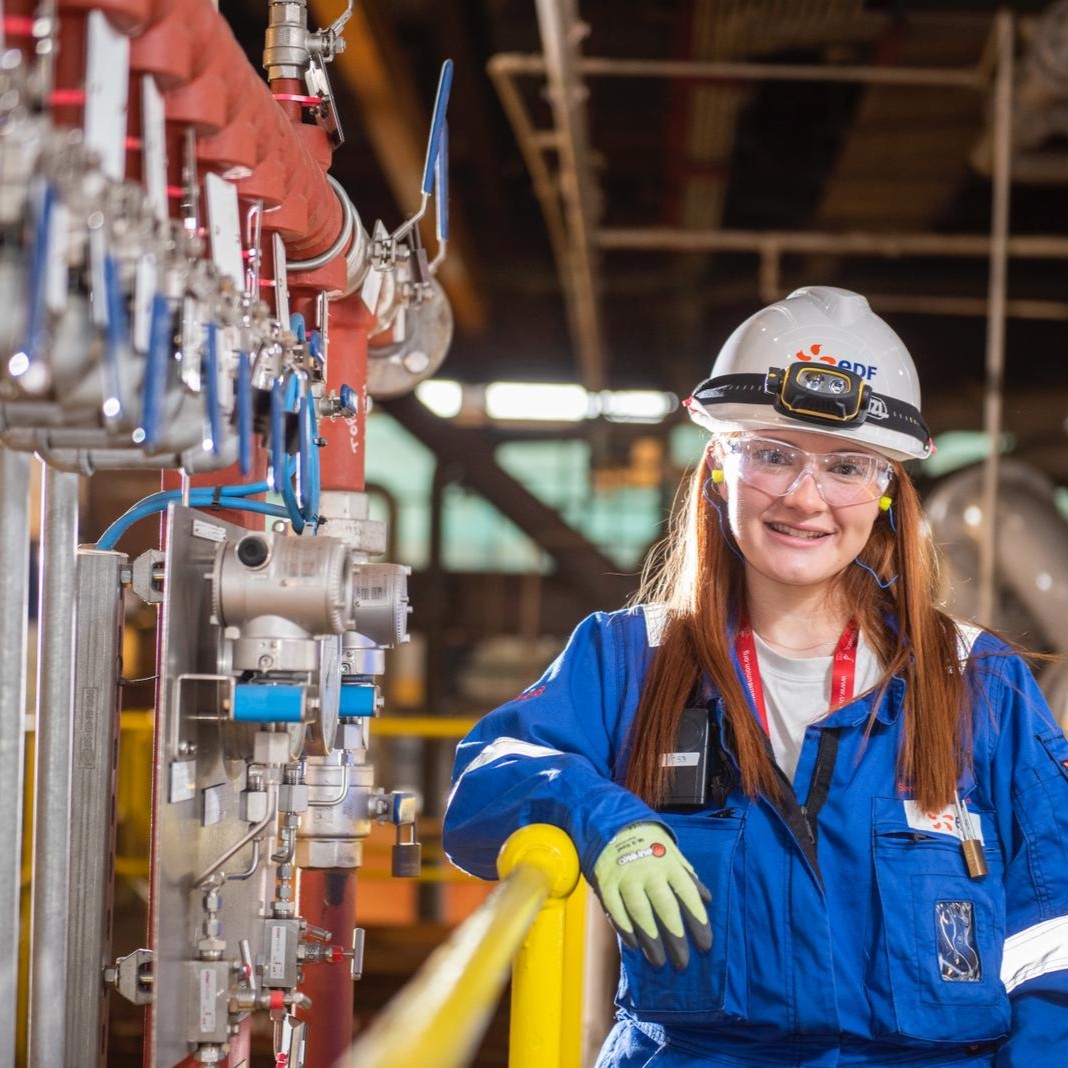 Siwan wearing protective overalls, gloves, safety glasses and a hard hat, leaning against a rail next to pipes.