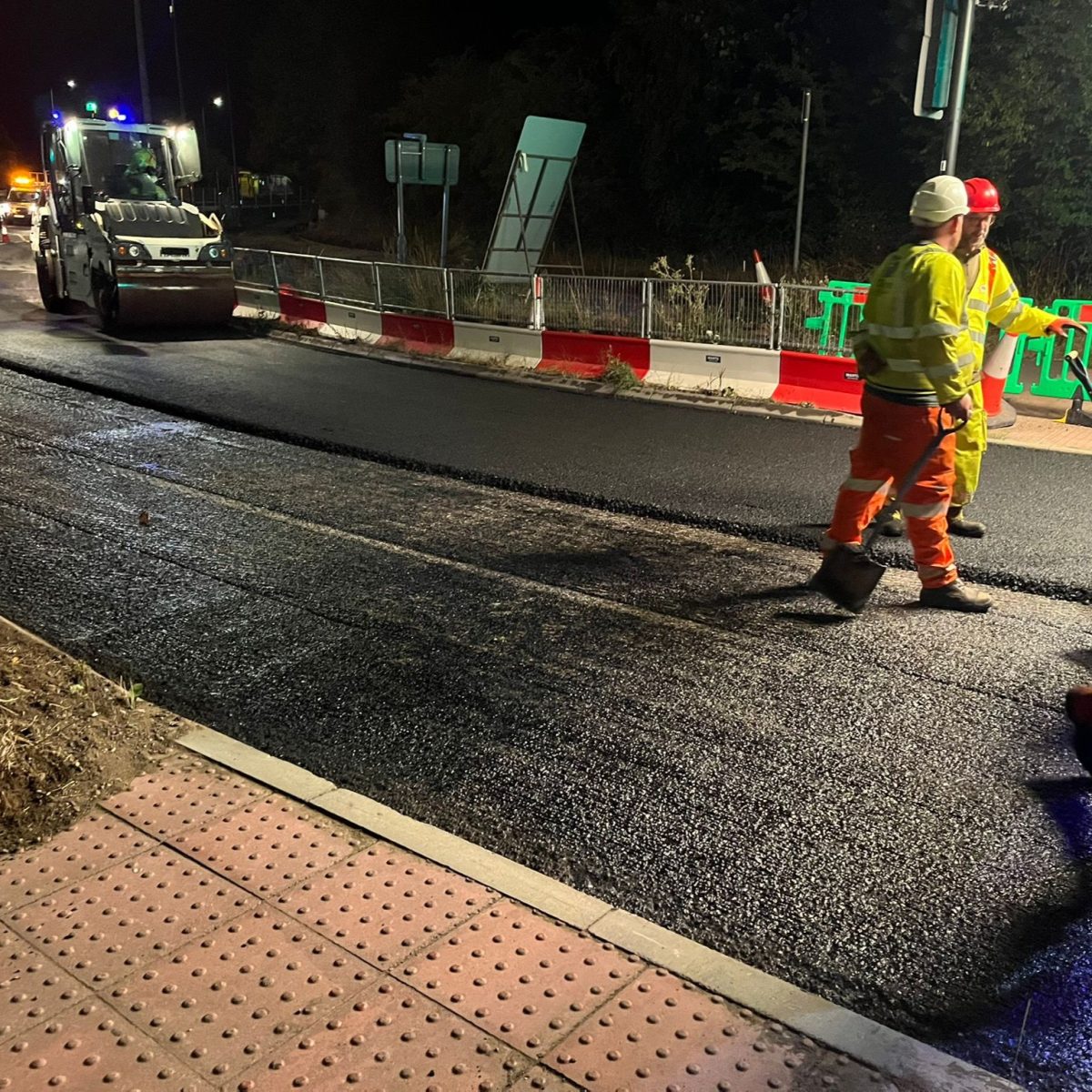 Workpeople wearing hi-vis outfits and hardhats talking during a road retarmacking at night.