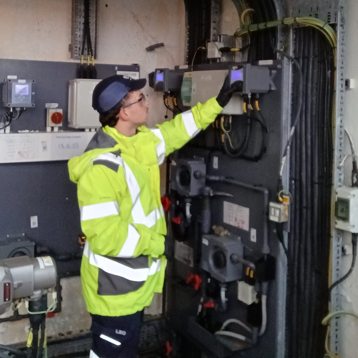 Water Quality Technician Bryce, wearing a hi-vis coat, gloves, and safety goggles, standing in a water processing facility, checking a piece of equipment fixed to the wall.