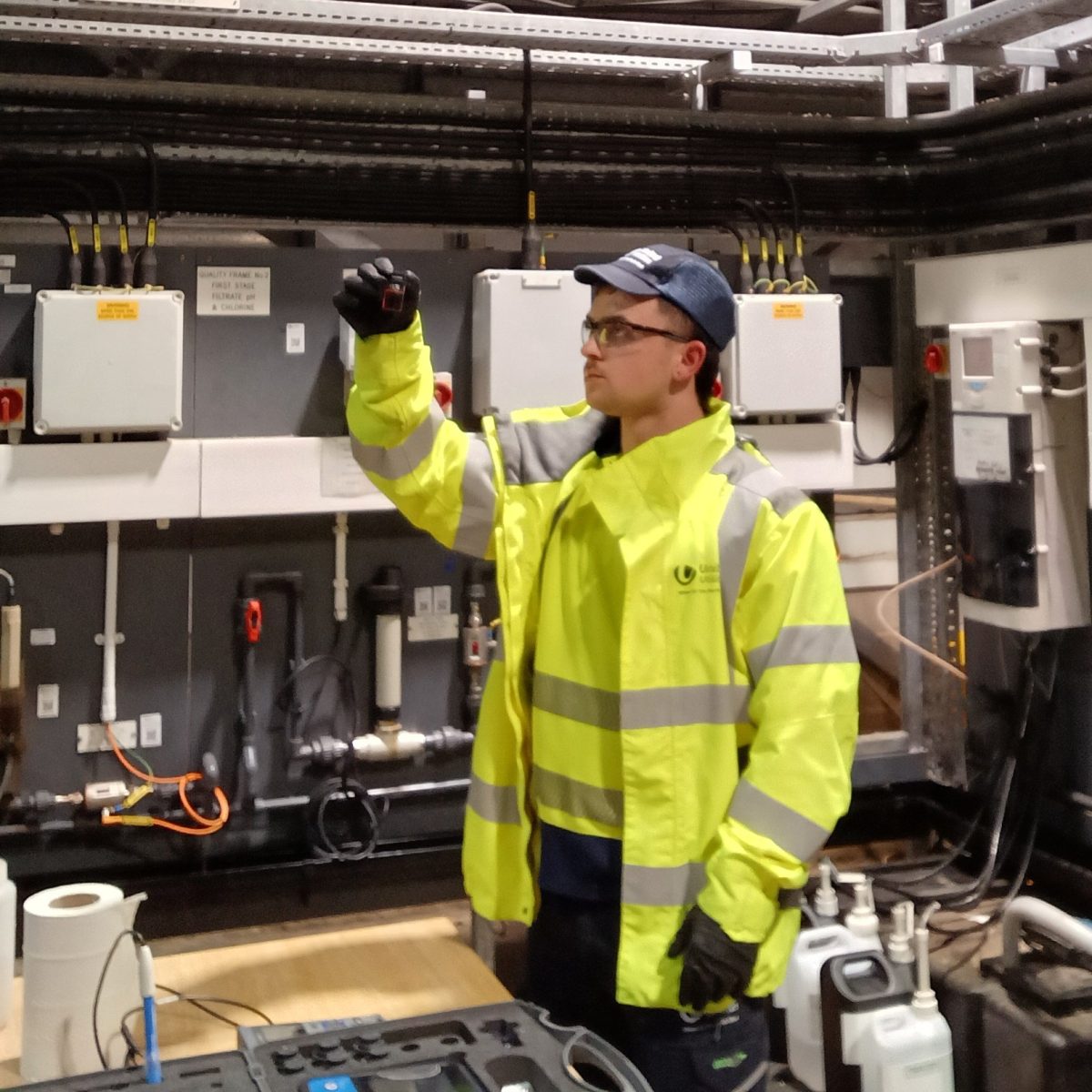 Water Quality Technician Bryce, wearing a hi-vis coat, gloves, and safety goggles, standing in a water processing facility, holding up a vial of liquid.
