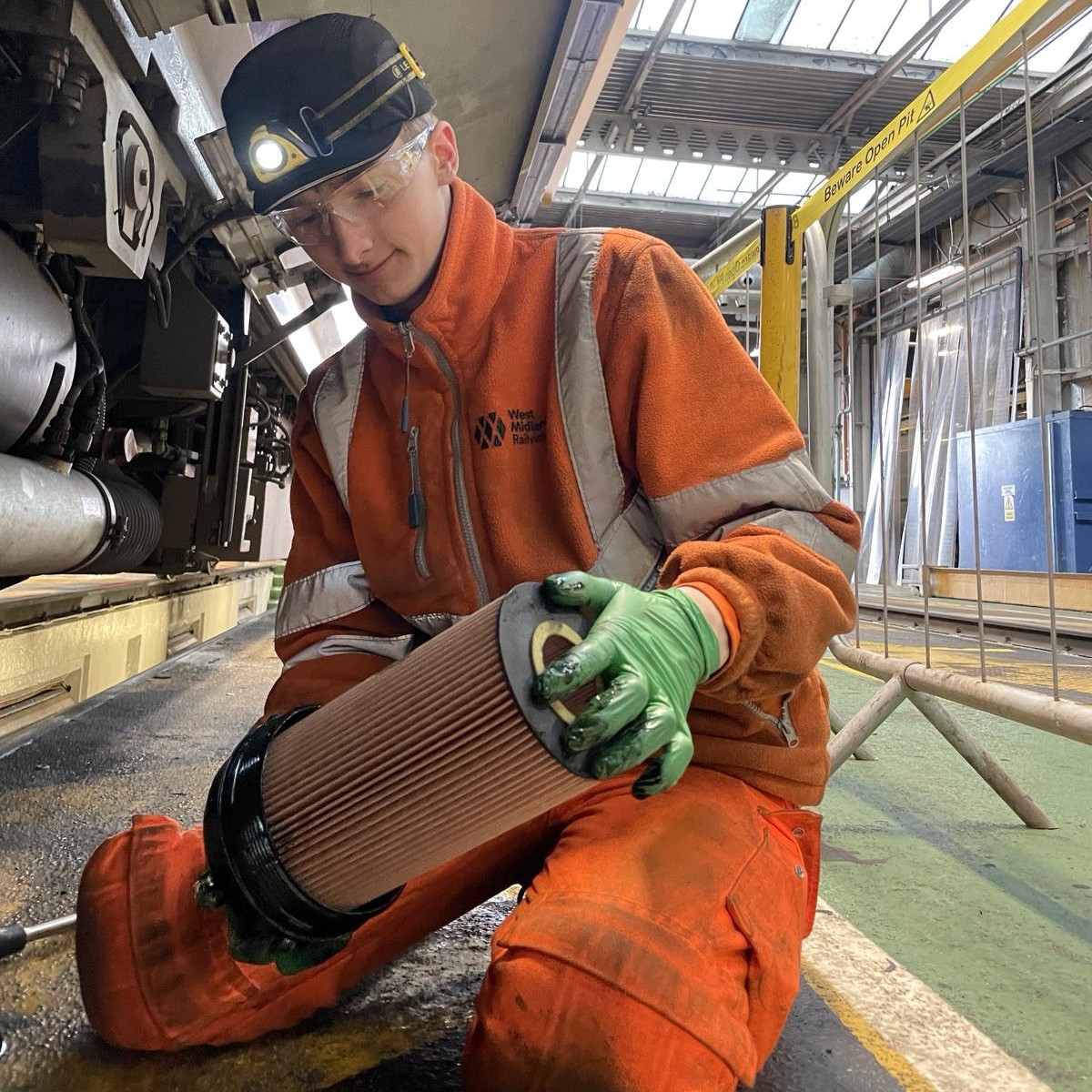 Jack, wearing overalls, gloves, safety goggles and a hard hat, kneeling beside a train, replacing a filter.