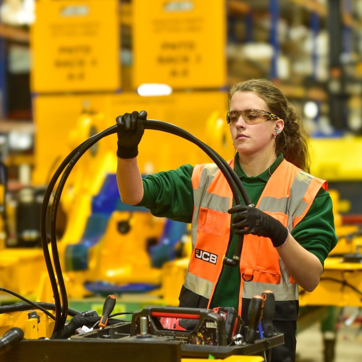 Kim the engineering and manufacturing technician, working on a machine in the JCB factory, wearing a hi-vis vest, gloves, and safety goggles.