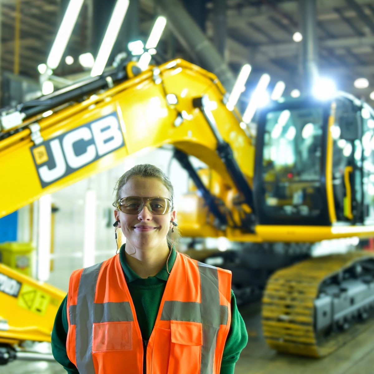 Kim the engineering manufacturing technician, wearing a hi-vis jacket and safety goggles, standing in front of a JCB excavator.