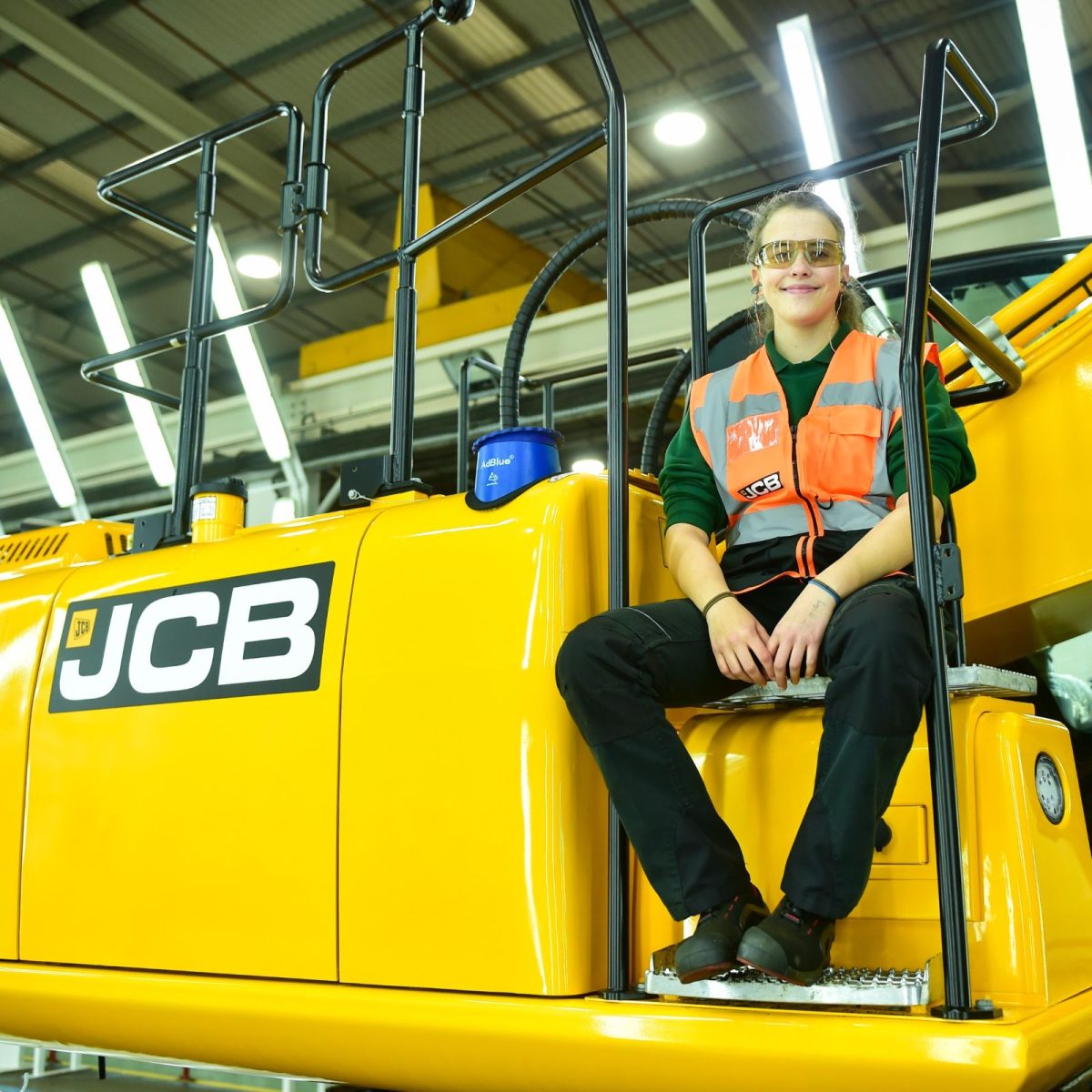 Kim the engineering manufacturing technician, wearing a hi-vis vest and safety goggles, sat on a JCB excavator.