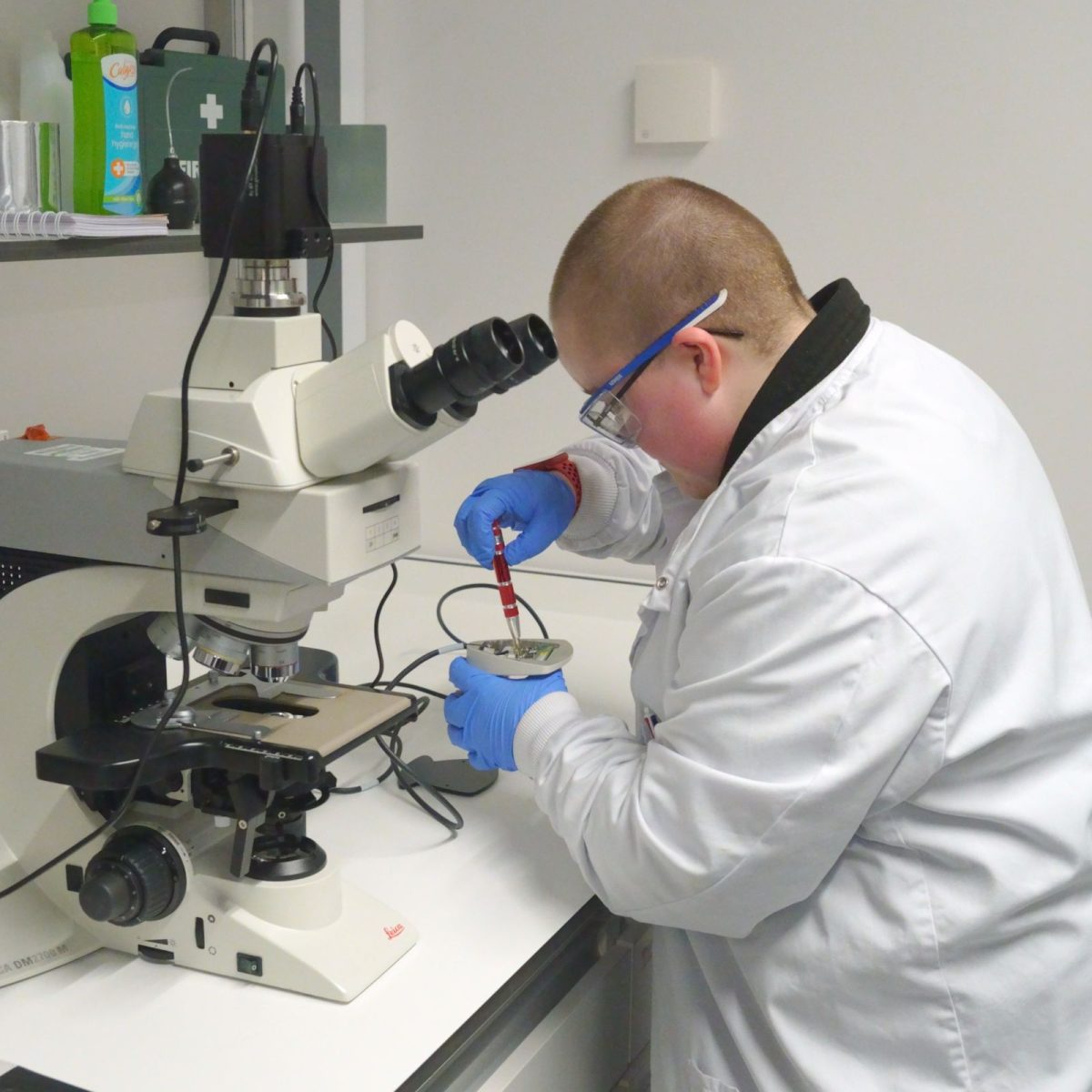Megan in the lab, wearing a lab coat, gloves and safety goggles, fixing an electrical component next to a microscope.