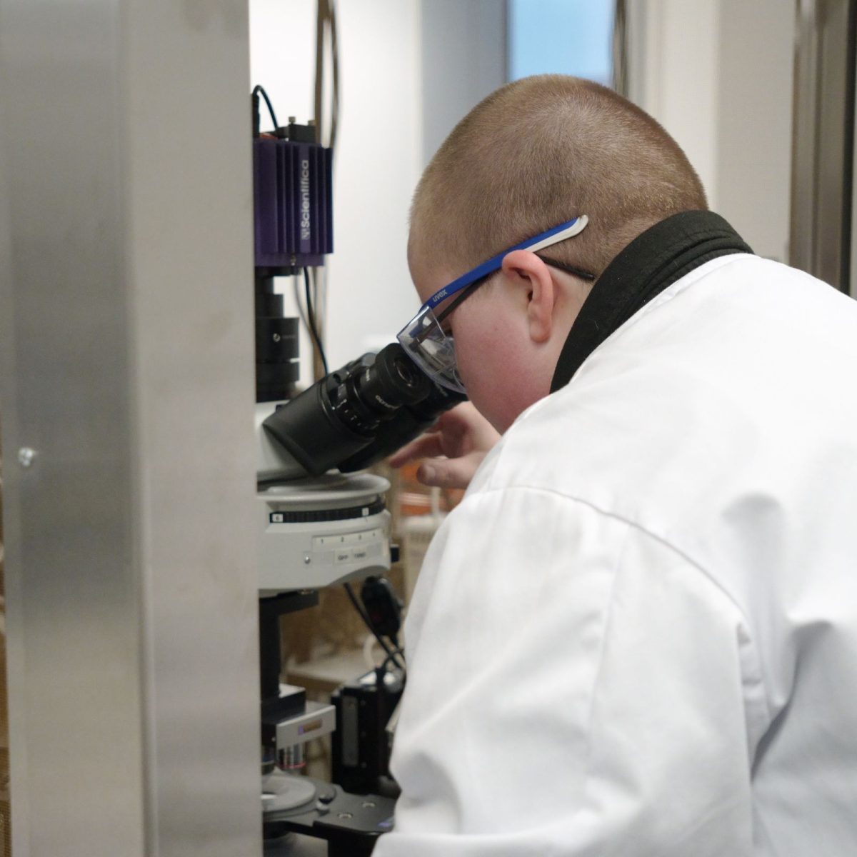 Megan in the lab, wearing a lab coat, looking down a microscope.