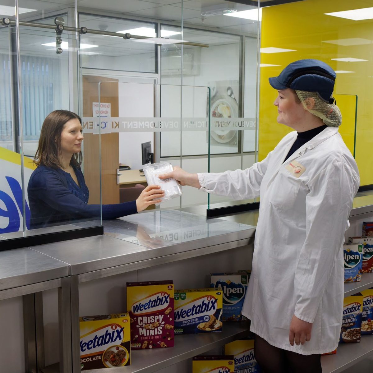 Phoebe wearing a lab coat, gloves, and cap, handing a trial product to a colleague over a counter.
