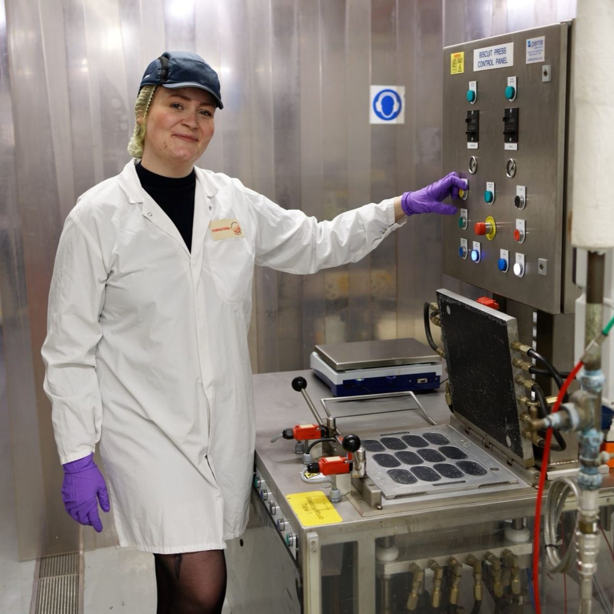 Phoebe wearing a lab coat, gloves, and cap, posing next to a piece of machinery in the test kitchen.