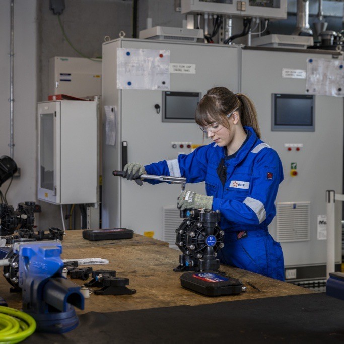 Maintenance and Operations Engineering Technician Anabella, wearing overalls, gloves and protective glasses, using a tool on a piece of equipment on a workbench.