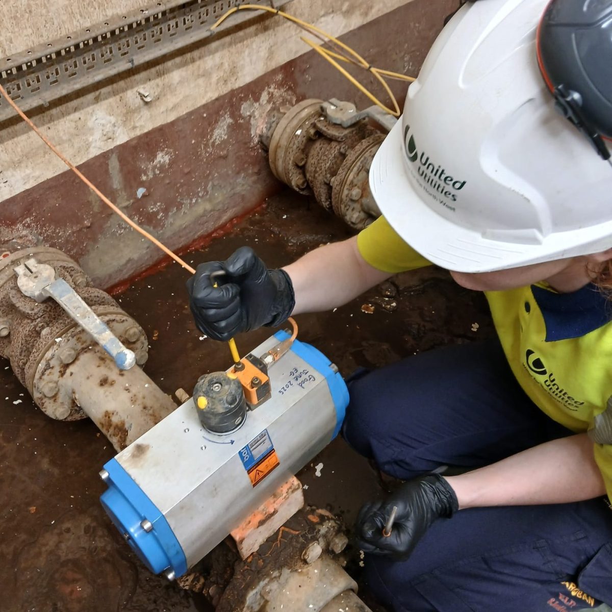 Water Process technician Ella, wearing a hard hat and gloves, crouching next to a water pipe, checking a pump.