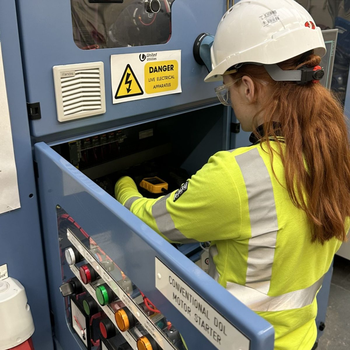 Water Process technician Ella, wearing a hard hat, gloves, and hi-vis top, checking equipment in an electrical control panel.