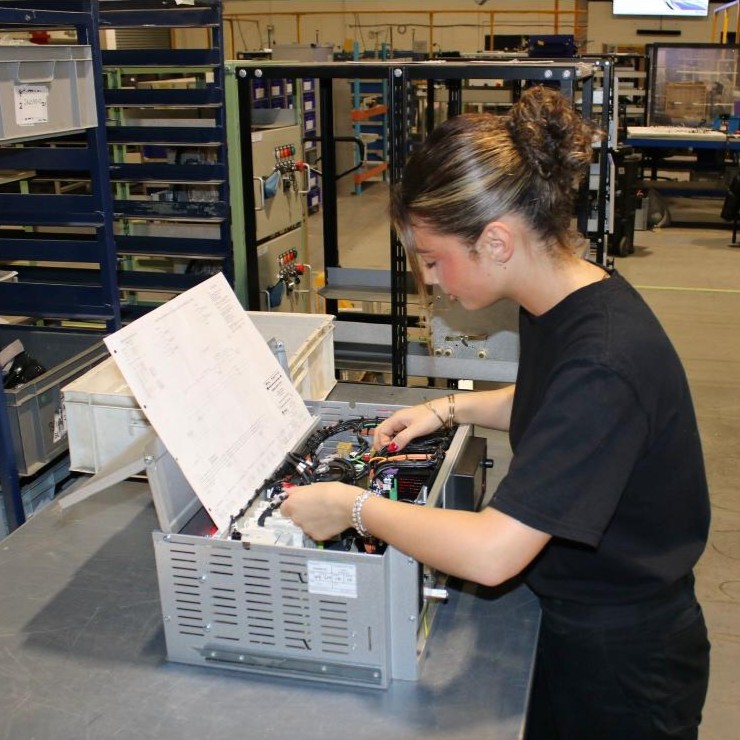 Engineering Fitter Imogen, on the workshop floor, following a wiring diagram to wire electrical components inside a box, on the tabletop.