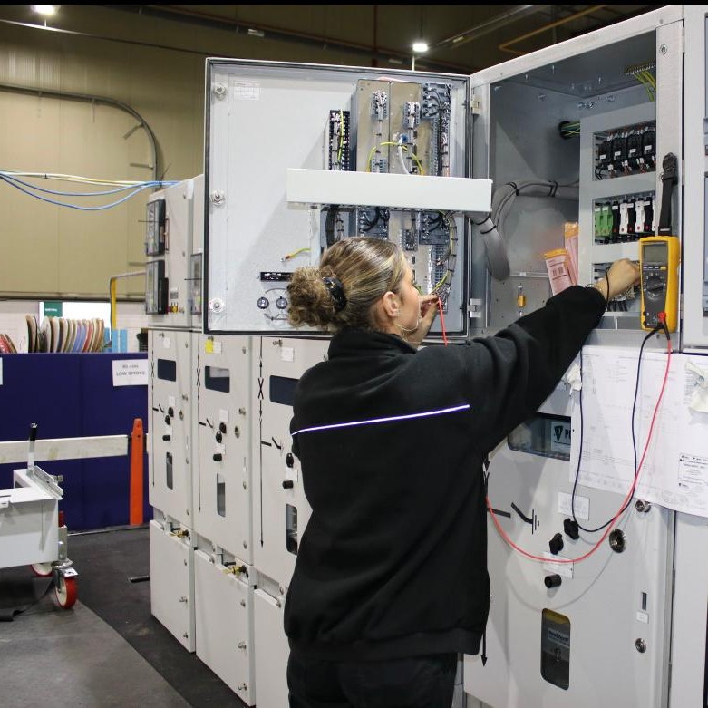 Engineering Fitter Imogen, on the workshop floor, wiring up electrical components in a cabinet, using a multimeter.