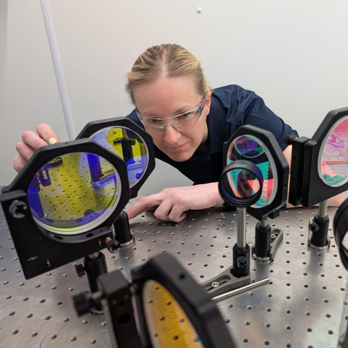 Research and development technician Kate, in the lab, wearing safety goggles, precisely aligning an array of lenses.